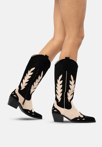 Pies que llevan botas vaqueras de gamuza negras y beige hasta la mitad de la pantorrilla, con patrones en forma de hoja y tacones bajos apilados, mostrados caminando sobre un fondo blanco.