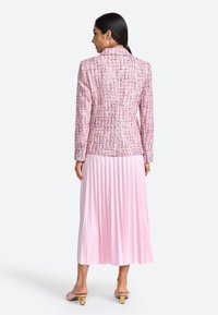 Blazer en tweed rose avec motif texturé, coupe ajustée et revers crantés, associé à une jupe plissée rose clair et des mules à talons rose pâle.