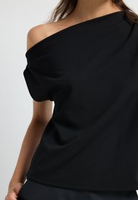 Top nero scollato sulla spalla realizzato in tessuto morbido, caratterizzato da una vestibilità relax e maniche corte, con una texture liscia e cuciture minime.
