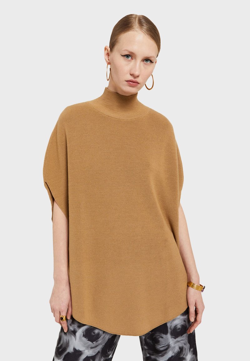 Ruskea ribattu poncho-neule, jossa on korkea kaulus, rento malli ja sivuaukot. Yhdistetty kuvioituihin housuihin ja rohkeisiin asusteisiin.