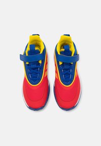 Baskets de sport rouges et bleues avec des accents jaunes, présentant une tige en mesh, un col rembourré et une sangle Velcro. Semelle en caoutchouc pour une meilleure adhérence.