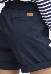 Mörkblå shorts med elastisk midja, två fickor och en läderlogopatch på baksidan. Tyget verkar lätt och slätt.