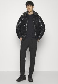 Chaqueta negra tipo puffer con acabado brillante y capucha, combinada con una camisa oscura y pantalones negros. El conjunto incluye calzado oscuro.