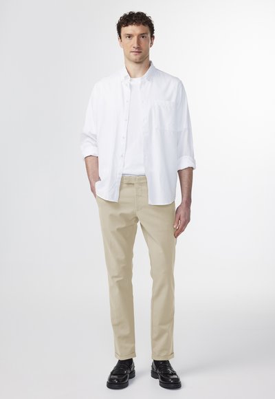 Mandens outfit består af en hvid skjorte med knapper, beige chinos og sorte sko. Skjorten har en lomme, og chinos har en lige pasform.