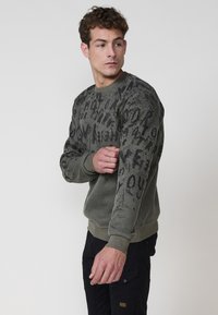Olívazöld pulóver, texturált kötött anyagból, fekete betűmintákkal, bordás mandzsettával és szegéllyel. A modell egy karját felemelve pózol.