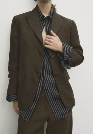 Persona che indossa una giacca e pantaloni marroni sopra una camicia a righe verticali nere e grigie, con la mano che sistema la patta della giacca.