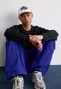 Camiseta negra de manga larga, pantalones sueltos azules, gorra blanca con texto negro y zapatillas grises. Postura sentada contra una pared.