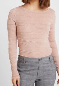 Ljust rosa långärmad tröja med texturerade horisontella ränder, kombinerad med grå rutiga byxor med knappstängning och fickor.