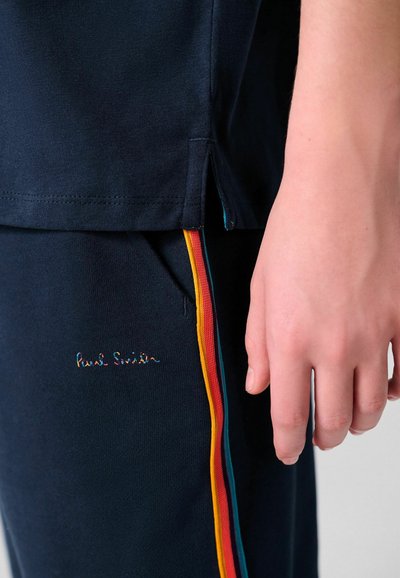 Mano apoyada junto a pantalones azul marino oscuro con franjas laterales en naranja, rojo y verde azulado, y camisa azul marino a juego con abertura lateral.