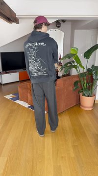 Sweatshirt negru cu glugă și design floral alb la spate, asortat cu pantaloni largi gri. Outfit casual într-un mediu de acasă, cu plante de interior.