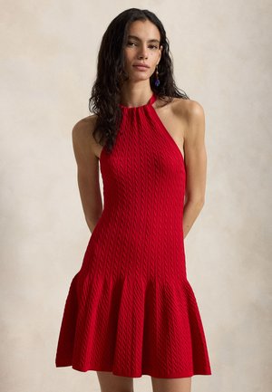 Γυναίκα με μακριά σκούρα σγουρά μαλλιά, ντυμένη με αμάνικο κόκκινο φόρεμα πλεκτό με σχέδιο καλώδιο και άνοιγμα στη φούστα, στέκεται μπροστά σε ουδέτερο φόντο.