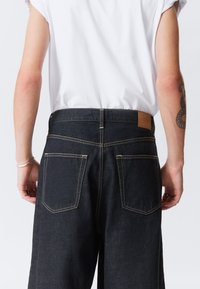 Persona vestida con pantalones vaqueros oscuros y una camiseta blanca, con un tatuaje visible en el brazo izquierdo, de pie contra un fondo claro y liso.