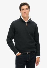 Maglione nero con colletto a zip, caratterizzato da una finitura testurizzata. Indossato sopra una camicia bianca con colletto, abbinato a jeans di denim scuri.