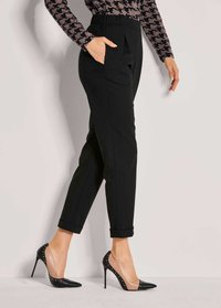 Zwarte op maat gemaakte broek met een geplooide voorkant en zijzakken, gecombineerd met zwarte en transparante hoge hakken met studs.