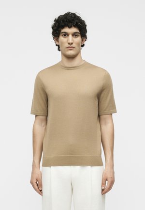 Άνδρας με σγουρά σκούρα μαλλιά, που φοράει μπεζ πουλόβερ με κοντά μανίκια και άσπρο παντελόνι, στέκεται μπροστά σε απλό φωτεινό φόντο, κοιτώντας μπροστά.