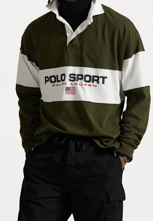 Osoba nosząca oliwkowo-białą koszulkę rugby z długim rękawem z napisem "Polo Sport Ralph Lauren" i amerykańską flagą, w połączeniu z czarnymi spodniami.