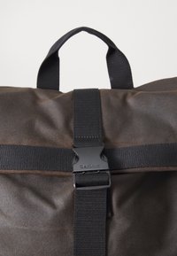 Close-up of brown fabric bag with black strap, black plastic buckle engraved with "Barbour," and black carry handle loop.