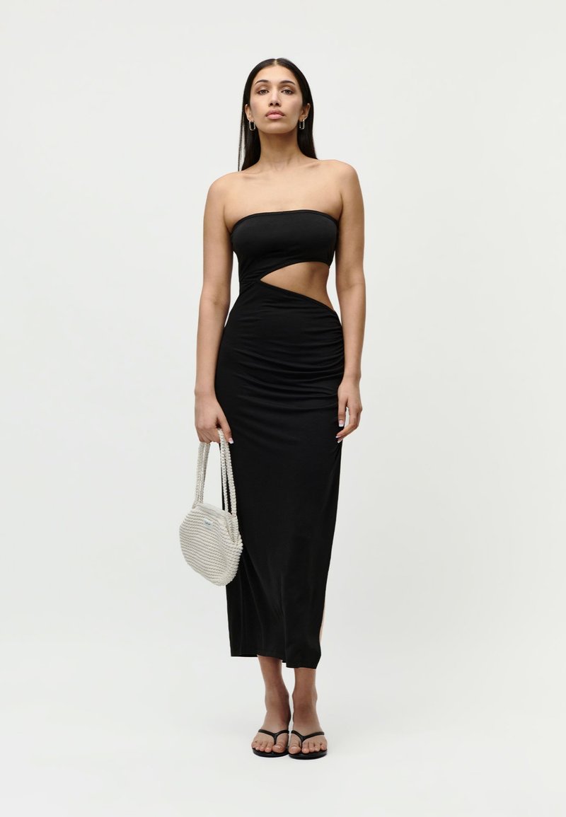 Γυναίκα που στέκεται φορώντας μαύρο strapless φόρεμα με πλευρικό άνοιγμα, κρατώντας μια λευκή πλεκτή τσάντα και φοράει μαύρα σανδάλια σε απλό φόντο.