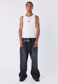 Camiseta blanca sin mangas con logo negro, combinada con jeans de mezclilla oscuros de tiro holgado y zapatillas negras, posando contra un fondo liso.