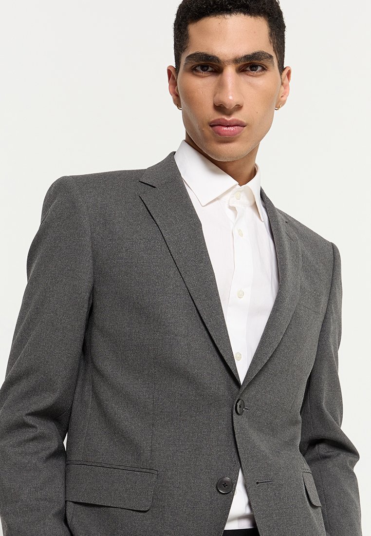 Blazer gris ajusté avec revers crantés, fermeture à un bouton et deux poches avant porté sur une chemise blanche à col. Texture lisse.