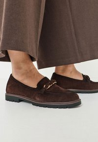 Braune Wildleder-Slipper mit geflochtenem Akzent, flacher schwarzer Gummisohle und Steppnaht-Details. Getragen zu einem dunklen braunen langen Rock.