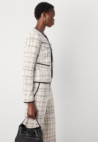 Veste en tweed texturé noire et blanche avec une coupe cintrée, présentant un bord et des poches noirs contrastants, assortie à un pantalon et un sac tissé noir.