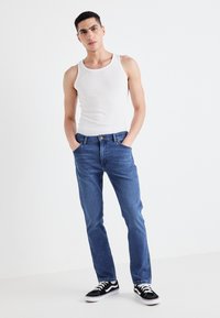 Biała prążkowana tank top, niebieskie dżinsy o prostym kroju i czarne sneakersy. Model stoi z rękami w kieszeniach na neutralnym tle.