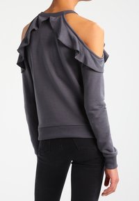 Sudadera gris con diseño de hombros al descubiertos, que presenta acentos con volantes en la parte posterior, puños acanalados y un tejido suave y texturizado.