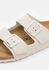 Sandale en suede beige clair avec deux sangles réglables dotées de boucles, une semelle en liège et une semelle extérieure en caoutchouc texturé.