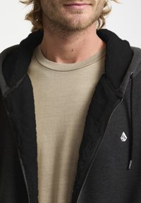 Hættejakke i sort med en fuzzy foring, der har en lys beige skjorte under. Bemærkelsesværdigt logo på jakken; blød tekstur generelt.