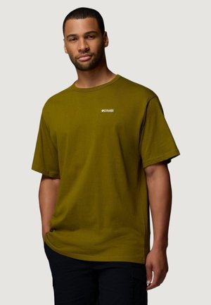 Uomo che indossa una maglietta Columbia verde oliva con logo sul petto e pantaloni neri, in piedi con una mano in tasca su uno sfondo semplice.