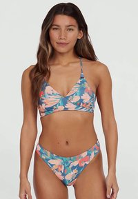 Γυναίκα φορεμένη με μπλε μπικίνι με ροζ και πορτοκαλί λουλούδια, σStanding με φόντο απλό ανοιχτό χρώμα, στραμμένη προς τα μπροστά.