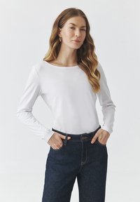 Camită albă cu mânecă lungă din material moale, cu un decolteu rotund, asortată cu pantaloni din denim de culoare bleu închis, cu închidere cu nasture și buzunare laterale.