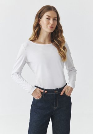 Camiseta de manga larga blanca de tejido suave con escote redondeado, combinada con pantalones vaqueros oscuros con cierre de botón y bolsillos laterales.
