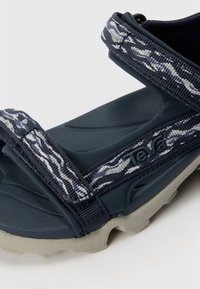 Mörkblå sandal med texturerad gummisula, nylonremmar med ett vitt och grått mönster och Teva-logotyp på sidan.