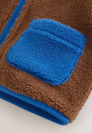 Material maro din fleece cu un buzunar din fleece albastru. Prezintă un fermoar albastru contrastant și o margine de material albastru neted în partea de jos.