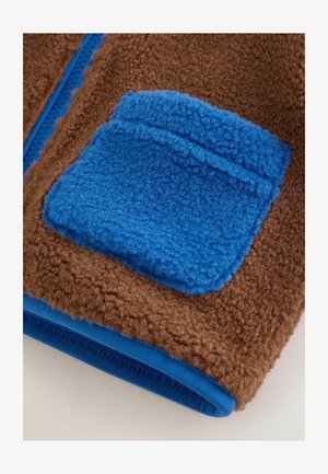 Brauner Fleece-Stoff mit einer blauen Fleece-Tasche. Verfügt über einen kontrastierenden blauen Reißverschluss und einen glatten blauen Stoffbesatz am unteren Rand.