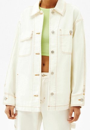 Personne portant une veste écrue boutonnée avec poches sur la poitrine et coutures claires, par-dessus un haut court en mesh vert et un pantalon écru assorti.