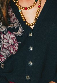 Cardigan noir avec broderie florale en bordeaux et or, doté de cinq boutons et de colliers superposés en or et perles colorées.