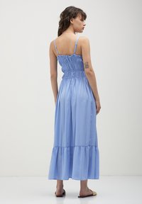 Frau mit welligem, dunklem Haar, die ein hellblaues, trägerloses Midikleid und geflochtene Rutschsandalen trägt, stehend vor einem schlichten weißen Hintergrund.