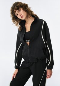 Giacca nera con zip e dettagli in bianco, realizzata in materiale leggero. Presenta un colletto alto e polsini elasticizzati, abbinata a capo sportivo nero.