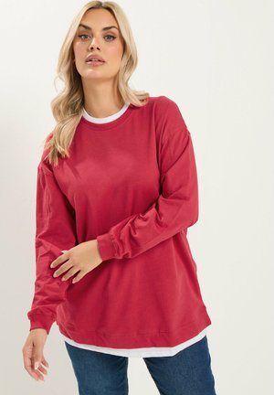 Mujer con cabello largo y rubio que lleva una sudadera roja holgada sobre una camisa blanca y vaqueros azules, de pie frente a un fondo claro y sencillo.