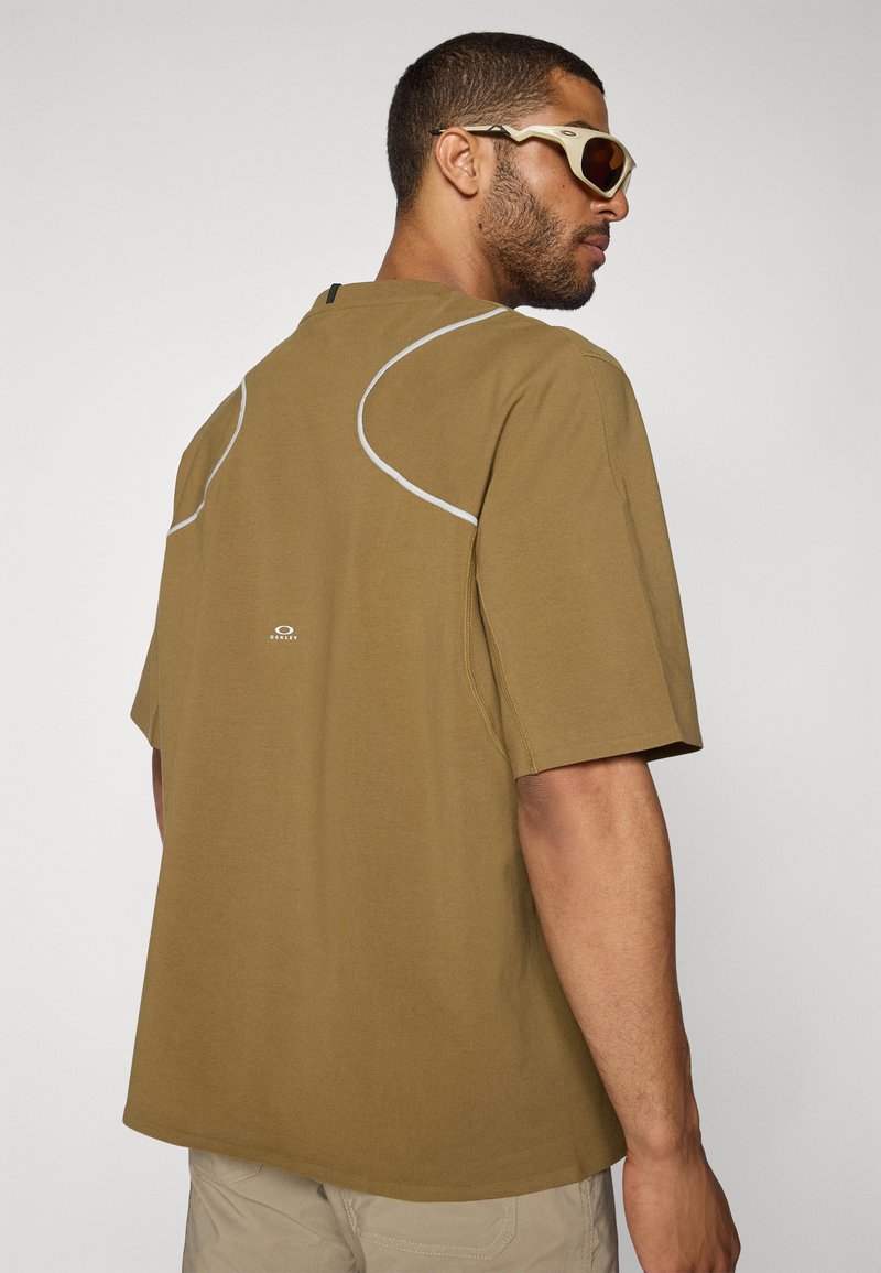 Brun kortärmad skjorta med reflekterande sömmar, rund halsringning och en logo på baksidan. Mjukt tyg med en avslappnad passform.