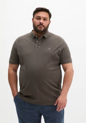 Mężczyzna z brodą w ciemnoszarej koszulce polo i niebieskich spodniach, stojący z rękami w kieszeniach na jednolitym białym tle.