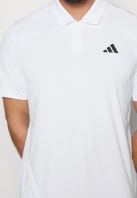 Polo blanc en tissu lisse, avec une patte de boutonnage à trois boutons et un logo Adidas noir sur le côté gauche de la poitrine. Panneaux latéraux texturés.