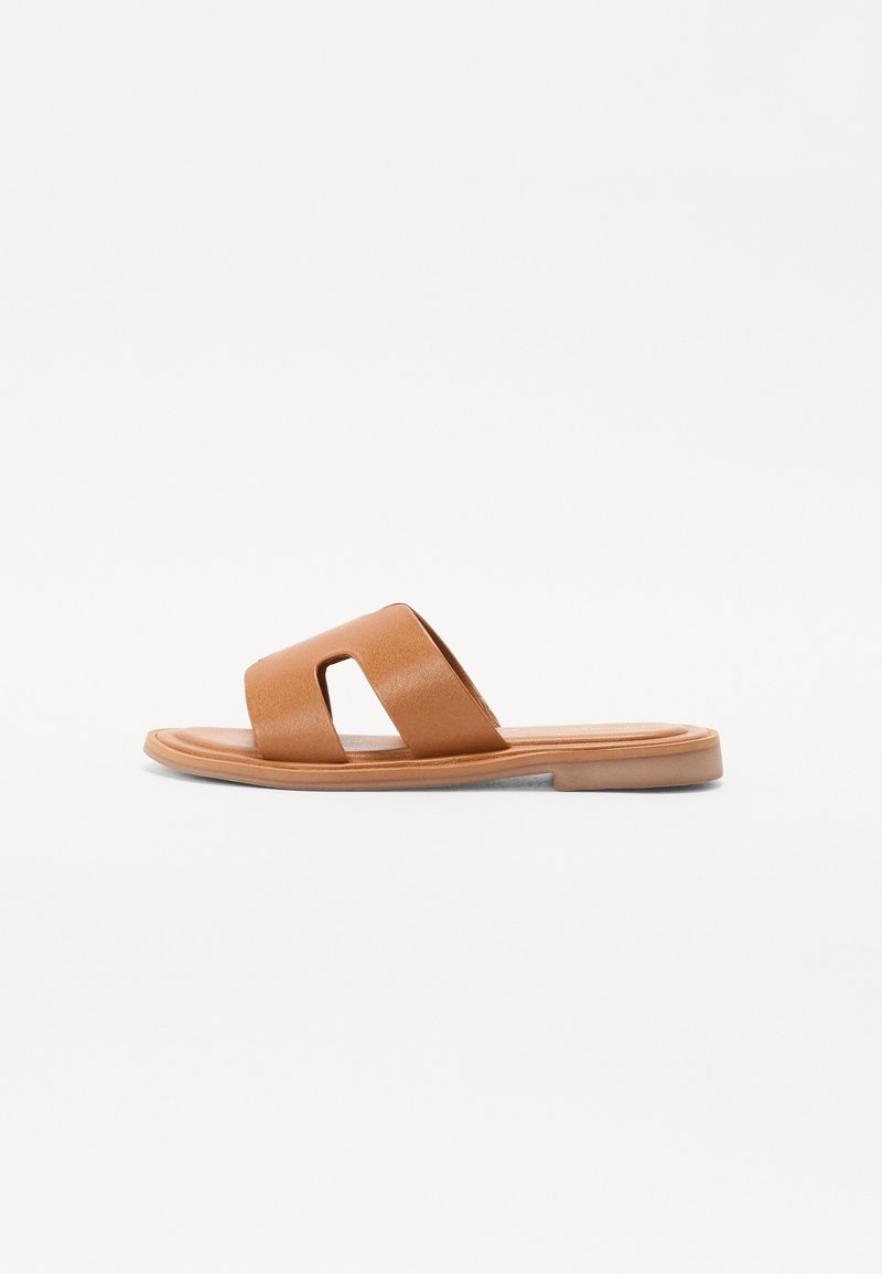 Brune skinnslidesandal med flat såle, med en bred stropp med utskjæringsdetaljer og en glatt tekstur. Enkel og minimalistisk design.