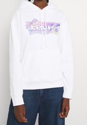 Biała bluza z kapturem Levi's z kolorowym pastelowym wzorem motyla i znakiem pokoju na przodzie, noszona z niebieskimi jeansami przez osobę o ciemnej karnacji.