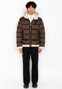 Brązowa kurtka puchowa z kremowym sztucznym futrem, zapinana na zamek z przodu, z dwoma bocznymi kieszeniami. Czarne akcenty i guziki na klatce piersiowej. Noszone czarne spodnie.
