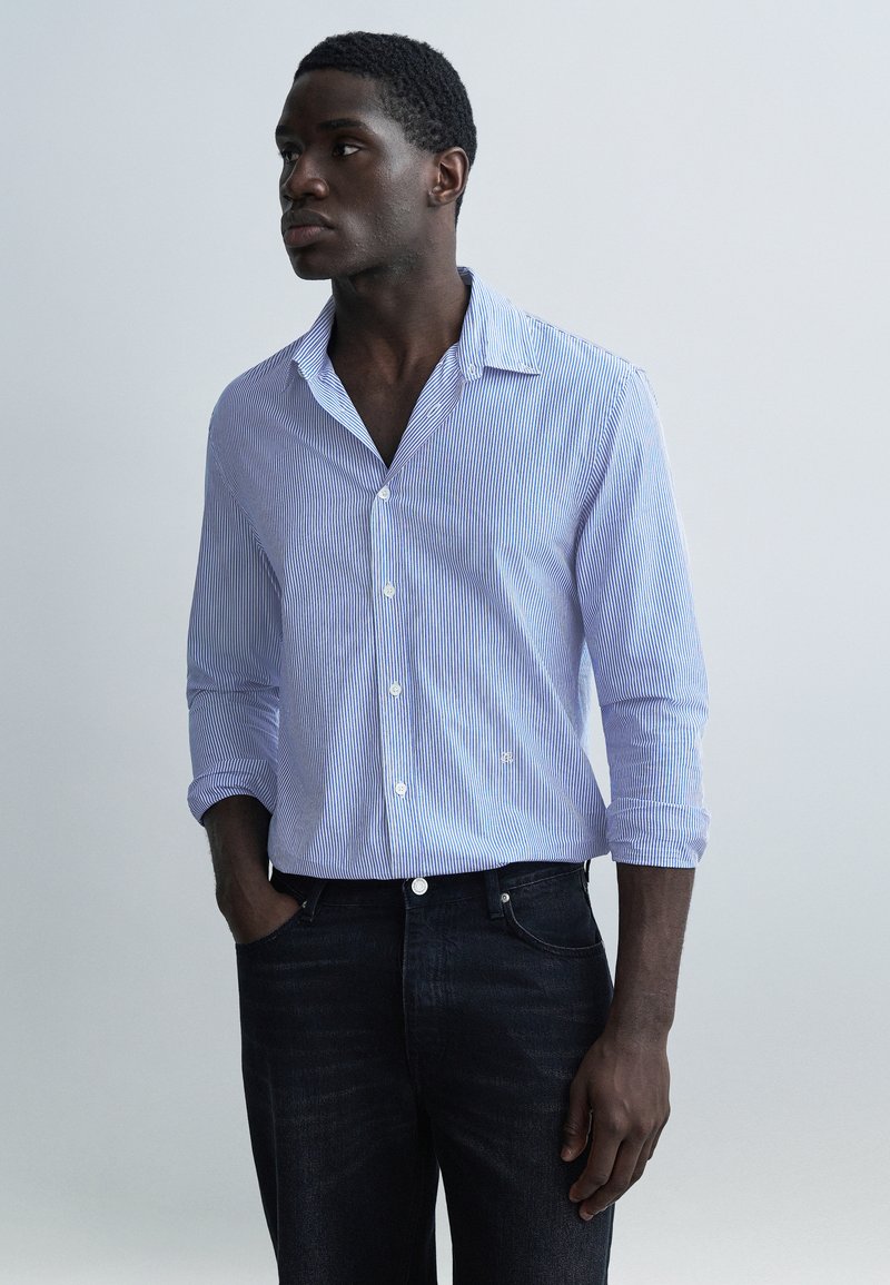 Lyseblå og hvide stribede skjorte med krave, lange ærmer rullet op, kombineret med mørke denimjeans. Model stående med den ene hånd i lommen.