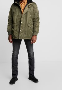 Doudoune vert olive avec une capuche à col noir, des boutons-pression, deux poches avant et des poignets côtelés, portée sur une chemise à carreaux et un jean foncé.